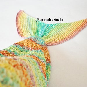 May include: A knitted mermaid tail blanket in vibrant colours of yellow, orange, green, and blue. The tail fin has a gradient of yellow, orange, and pink. The text "@annaluciadu" is visible.