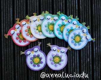May include: A collection of colorful, crocheted owl-shaped coasters. Each coaster features a white base with a central floral design in various colors, including yellow, orange, green, and blue. The owls have closed eyes and small tufts of yarn. The background is a dark wooden surface.