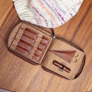 Op de afbeelding: Een bruin lederen etui met ritssluiting bevat breibenodigdheden, waaronder diverse metalen rondbreinaaldpunten, een driehoekig lederen tasje voor een schaar en een donkerhouten gereedschap, naast een wit gebreid item.