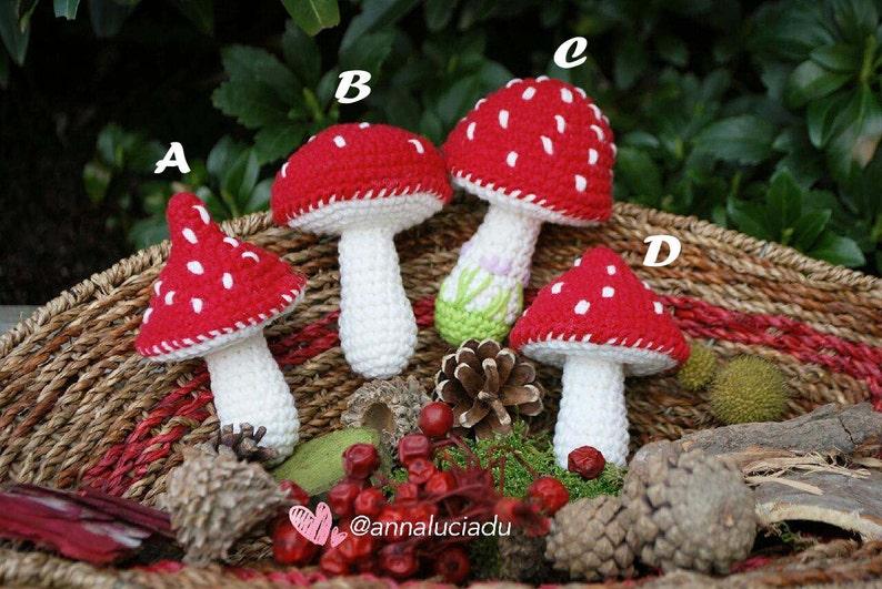 May include: Four crocheted red and white mushroom toys with white stems in a wicker basket with green moss, pine cones, and red berries. The mushrooms are labeled A, B, C, and D.