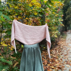 May include: A pink knitted shawl draped over a person's shoulders in a woodland setting. The shawl is made of a soft, fuzzy yarn and has a delicate, flowing drape. The person is wearing a green dress and is standing on a path surrounded by fallen leaves.