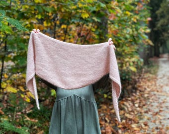 May include: A pink knitted shawl draped over a person's shoulders in a forest setting. The shawl is made of a soft, fuzzy yarn and has a delicate, flowing drape. The person is wearing a green dress and is standing in a path surrounded by fallen leaves.
