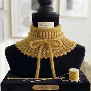 Op de afbeelding: Een mosterdgele gebreide kraag met een geribbelde textuur en een strik in het midden. Een klein wit label vermeldt Annalucia's Collar. Daaronder liggen een bijpassende garenklos en twee breinaalden op een zwarte voet met het opschrift Cashmere & Silk Blend.