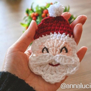 May include: A crocheted Santa Claus face ornament. The ornament features a red hat with a white pom-pom, a white beard, and a smiling face with black eyes and a red mouth. The ornament is handmade.