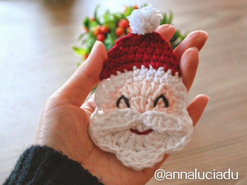 May include: A crocheted Santa Claus face ornament. The ornament features a red hat with a white pom-pom, a white beard, and a smiling face with black eyes and a red mouth. The ornament is handmade.