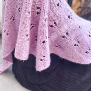 May include: A lilac knitted shawl featuring a floral pattern. The shawl is draped over a dark garment, highlighting the intricate openwork design. The soft, textured fabric creates a cosy and elegant look.