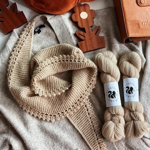 以下が含まれることがあります： ベージュ色のスカラップエッジのニットスカーフと、お揃いの帽子が、ライトグレーのカーディガンに展示されています。「Annalucia Design」のラベルが付いた2つの糸玉がスカーフの隣にあります。木製の花の形をした装飾と茶色の革のノートも表示されています。