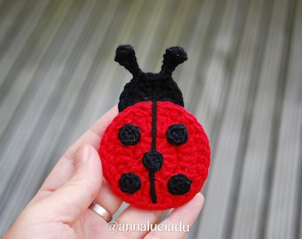 May include: A crocheted ladybug with a red body, black spots, and black antennae. The ladybug is held in a hand.