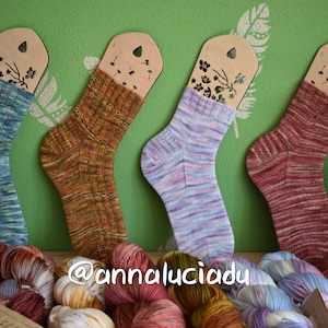 May include: Four pairs of hand-knitted socks displayed on wooden tags with floral designs. The socks are in various colors, including blue, orange, pink, and red. The socks are surrounded by balls of yarn in various colors.