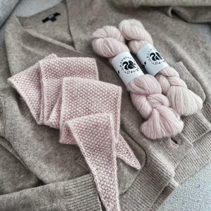 Puede incluir: Una bufanda de punto rosa con un extremo puntiagudo, un cárdigan gris y dos ovillos de hilo rosa con etiquetas que dicen "AnnaLu Design".