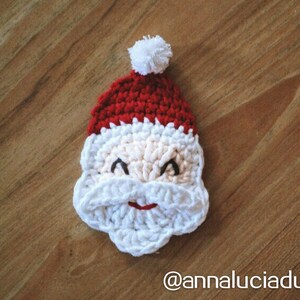 May include: A crocheted Santa Claus face with a red hat, white beard, and a small white pom-pom. The face is beige with black eyes and a red smile. The image is set against a wooden background.