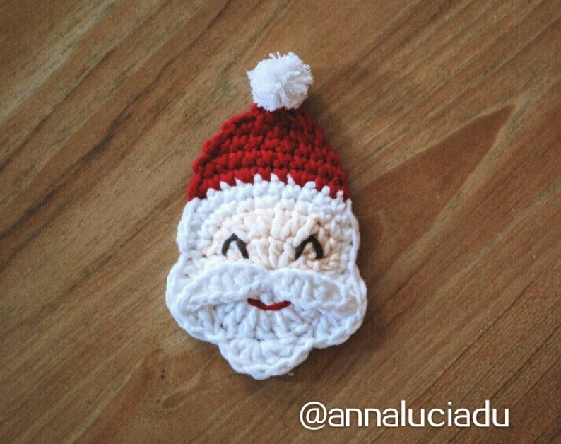May include: A crocheted Santa Claus face with a red hat, white beard, and a small white pom-pom. The face is beige with black eyes and a red smile. The image is set against a wooden background.