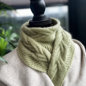 May include: A green knitted scarf with a cable knit pattern. The scarf is draped over a mannequin's neck.