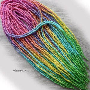 May include: Rainbow ombre wool dreadlocks, a type of synthetic hair extension, in shades of pink, purple, orange, yellow, and green. The dreadlocks are arranged in a loose pile on a wooden surface.