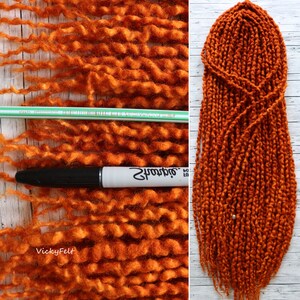 May include: A close-up of a set of long, orange-brown wool dreadlocks. The dreadlocks are made of thick, curly wool and are arranged in a neat, symmetrical pattern. The dreadlocks are shown against a white wooden background.