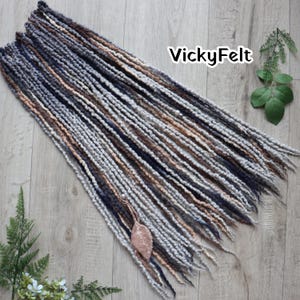 May include: A collection of handmade dreadlocks in various shades of gray, brown, and navy blue. The dreadlocks are arranged on a wooden surface, with the text "VickyFelt" visible. A decorative leaf charm is attached to the dreadlocks.