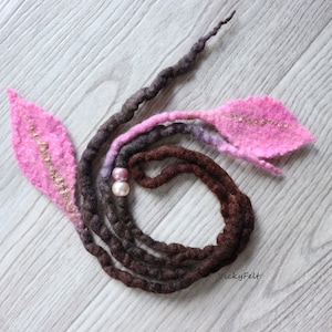 May include: Handmade felt hair accessory with a spiral of brown, purple, and pink dreadlocks. The design includes two pink leaf-shaped accents with gold stitching and two pink pearl beads. The accessory is designed for hair styling.