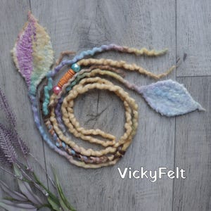 May include: A handcrafted hair accessory made from felt, featuring a leaf design. It includes a spiral of beige and brown wool dreadlocks, with pastel-colored strands and decorative beads. The text "VickyFelt" is visible.