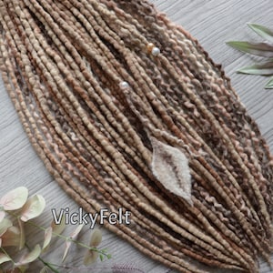 May include: A collection of handmade dreadlocks in various shades of brown and beige, with a few strands of pink. The dreadlocks are adorned with small pearl beads and a felt leaf. The text "VickyFelt" is visible.