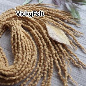 May include: Close-up of a bundle of light brown dreadlocks. The dreadlocks are thick and textured, with a wavy appearance. A light-colored leaf-shaped embellishment is attached to the dreadlocks. The text "VickyFelt" is displayed in white letters.