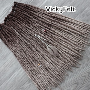 May include: Brown and tan ombre dreadlocks with the text "VickyFelt" in white. The dreadlocks are arranged in a layered pattern, with a few strands adorned with decorative beads and a feather-like embellishment. The overall colour scheme is a blend of brown and tan.