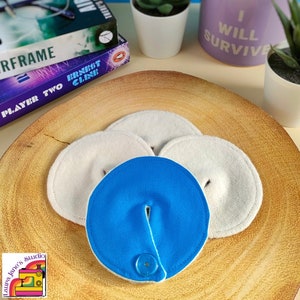 May include: Three white reusable breast pads with a blue reusable breast pad with a snap closure in the centre. The pads are on a wooden surface.
