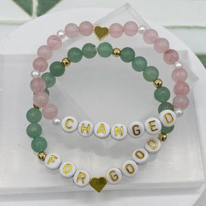 May include: Two beaded bracelets with gold accents. The top bracelet is pink and white with a gold heart charm. The bottom bracelet is green and white with gold letters spelling "CHANGED FOR GOOD".