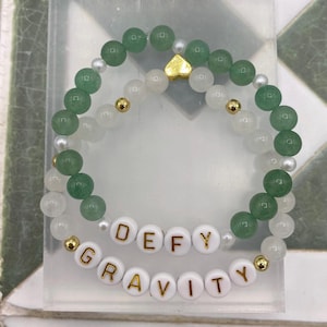 May include: A green and white beaded bracelet with a gold heart charm and white letter beads spelling "DEFY GRAVITY".