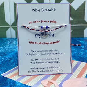Könnte beinhalten: Eine weiße Karte mit blauen und roten Schnurbändern, die um sie herum gebunden sind. Die Karte trägt den Text "Wish Bracelet" und ein Zitat über Träume, Magie und Wunder. Die Armbänder haben einen silbernen Anker-Anhänger.