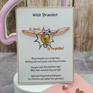 May include: A white card with the text "Wish Bracelet" in black lettering. The card features a colourful string bracelet with a key charm and a lightbulb graphic. The text on the card reads: "A Spark... Of Imagination! These bracelets are a simple token. Yet they hold much power when they are broken. Say your wish; then hold them tight. Wear them close both day and night. And when they break and fall apart, You'll find the wish spoken from your heart."