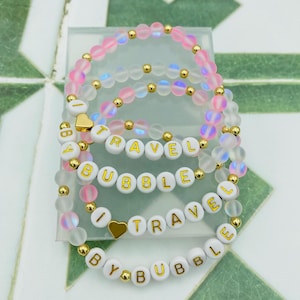 以下が含まれることがあります： 「I TRAVEL BY BUBBLE」の文字が金色の文字で書かれた白いビーズのブレスレットのセット。ピンク、クリア、ゴールドのビーズと、ゴールドのハートチャームが特徴です。