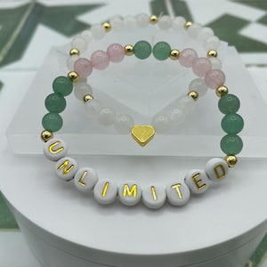 May include: Two bracelets, one with green and white beads and the other with pink and white beads. The green and white bracelet has gold beads and the word "UNLIMITED" spelled out in gold letters on white beads.