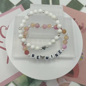 May include: Two beaded bracelets with pink, white and orange beads. The smaller bracelet has the words "BE MINE" spelled out with silver letter beads and a silver heart charm.