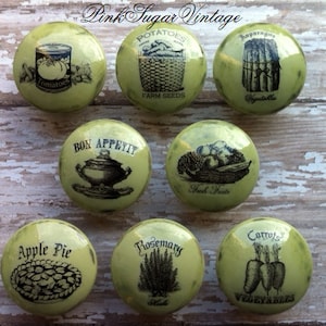 Peut inclure: Neuf boutons ronds en c&eacute;ramique verte avec des illustrations vintage en noir et blanc de nourriture et de l&eacute;gumes. Les boutons comportent des textes tels que "Tomates", "Pommes de terre", "Asperges", "Bon app&eacute;tit", "Fruits frais", "Tarte aux pommes", "Romarin" et "Carottes".