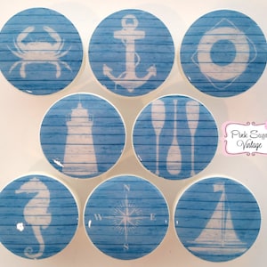 Può includere: Set di otto pomelli in ceramica bianca con disegni nautici blu e bianchi. I disegni includono un granchio, un'ancora, un salvagente, un faro, remi, una bussola, un cavalluccio marino e una barca a vela.