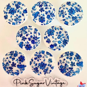 以下が含まれることがあります： 白地に青色の花柄が描かれた丸い白いセラミックタイルが8枚。タイルは白い表面に円形に配置されています。画像の下中央には「Pink Sugar Vintage」という文字が見えます。
