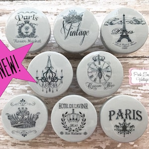 Może przedstawiać: Zestaw ośmiu szarych ceramicznych gałek z czarno-białymi wzorami w stylu vintage paryskim. Wzory obejmują koronę, pszczołę, zegar, żyrandol, wieżę Eiffla oraz słowa "Paryż", "Vintage" i "Queen Bee".