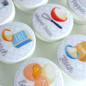 May include: Set of 10 white ceramic knobs with colorful illustrations of baking ingredients, including salt, sugar, flour, starch, whipped cream, baking powder, eggs, and pepper. Each knob features a different ingredient and the name of the ingredient is written below the illustration.