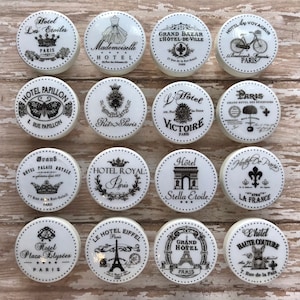 Op de afbeelding: Set van 12 witte keramische knoppen met zwart-witte Parijse ontwerpen. Elke knop heeft een ander Parijse monument, hotel of symbool, zoals de Eiffeltoren, de Arc de Triomphe en het Ritz Paris.