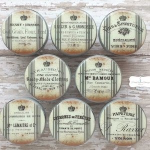 以下が含まれることがあります： 白とグレーのストライプの背景に、ヴィンテージスタイルの丸いキャビネットノブが8個セットになっています。各ノブには、黒と金色のテキストで、フランス風のデザインが施されています。テキストには、「Vins & Spiritueux」、「Ready-Made Clothing」、「Papeterie」など、企業名や製品名が含まれています。
