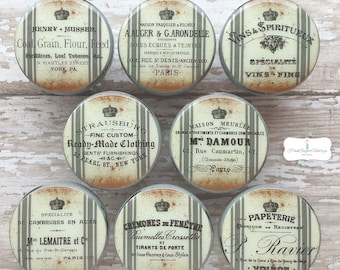 May include: Set of 8 vintage-style round cabinet knobs with a distressed white and grey striped background. Each knob features a different French-inspired design with text in black and gold. The text includes the names of businesses and products, such as "Vins & Spiritueux", "Ready-Made Clothing", and "Papeterie".