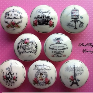 May include: Seven white ceramic knobs with vintage Parisian-themed designs. The designs include a birdcage, a crown, a chandelier, a tower, and text in French.