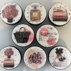 May include: Set of 8 round, black and white ceramic knobs with various designs. The designs include a bouquet of pink roses, a bottle of perfume, a stack of books with the text "Fashion in good taste" and "Boss Babe", a black handbag, a pink heart with the text "girl boss", a stack of books with the text "Fashion in good taste" and "Boss Babe", and a pair of black high heels.