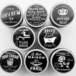 May include: A collection of vintage-style black and white tins with various labels. The labels include text such as "Soap & Water," "Olive Oil Soft Soap," "Powder Soap," "Hot Bath," "Cold Cream," and "Rose de Provence." The tins are arranged in a circular pattern.