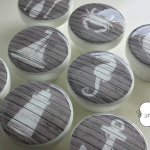 Peut inclure: Ensemble de huit boutons en céramique blanche sur le thème nautique. Chaque bouton présente un motif différent, notamment une boussole, un voilier, un phare, une ancre, un crabe, un hippocampe, une bouée de sauvetage et des rames. Les boutons ont un fond en bois gris.