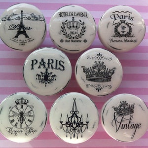 Op de afbeelding: Set van acht witte keramische knoppen met zwart-witte ontwerpen. De ontwerpen omvatten een kroon, een bij, een kroonluchter en het woord "Paris".