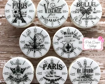 May include: Set of 8 white ceramic clock knobs with black and white vintage Parisian designs. The knobs feature various Parisian landmarks, including the Eiffel Tower, the Grand Palais, and the Jardin du Luxembourg. Each knob has a Roman numeral clock face and a unique design.