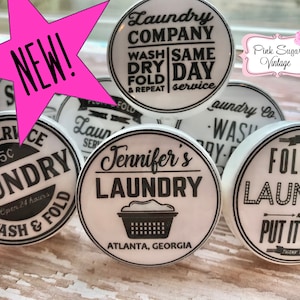 Peut inclure: Six boutons ronds en céramique blanche avec des inscriptions noires. Les boutons présentent différents motifs sur le thème de la lessive, notamment "Laundry Company", "Self Service Laundry", "Jennifer's Laundry" et "Fold the Laundry and Put it Away".