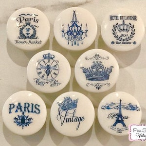 Może przedstawiać: Zestaw ośmiu białych ceramicznych gałek z niebieskimi i czarnymi wzorami. Wzory obejmują koronę, pszczołę, żyrandol, zegar, wieżę Eiffla oraz słowa "Paryż", "Vintage" i "Hotel de L'avenir".