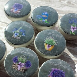 K&ouml;nnte beinhalten: Acht Keramikkn&ouml;pfe mit grauem Hintergrund und einem Lavendel-Design. Die Kn&ouml;pfe zeigen verschiedene Lavendelmotive, darunter Str&auml;u&szlig;e, K&ouml;rbe und Topfpflanzen. Der Text "LES JARDIN DE PROVENCE" ist auf jedem Knopf aufgedruckt.